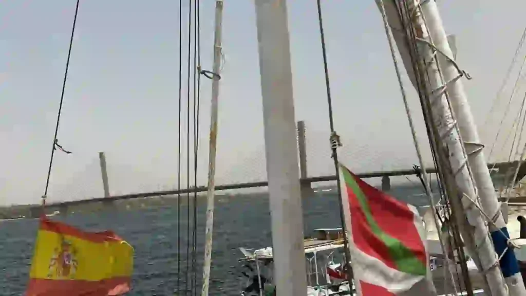 View from the deck of the felucca Maitea, with the Spanish and Basque flags fluttering above the Nile River, and the Aswan Bridge visible in the background.