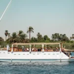 Traditional white dahabiya sailboat cruising on the Nile River with palm trees in the background.