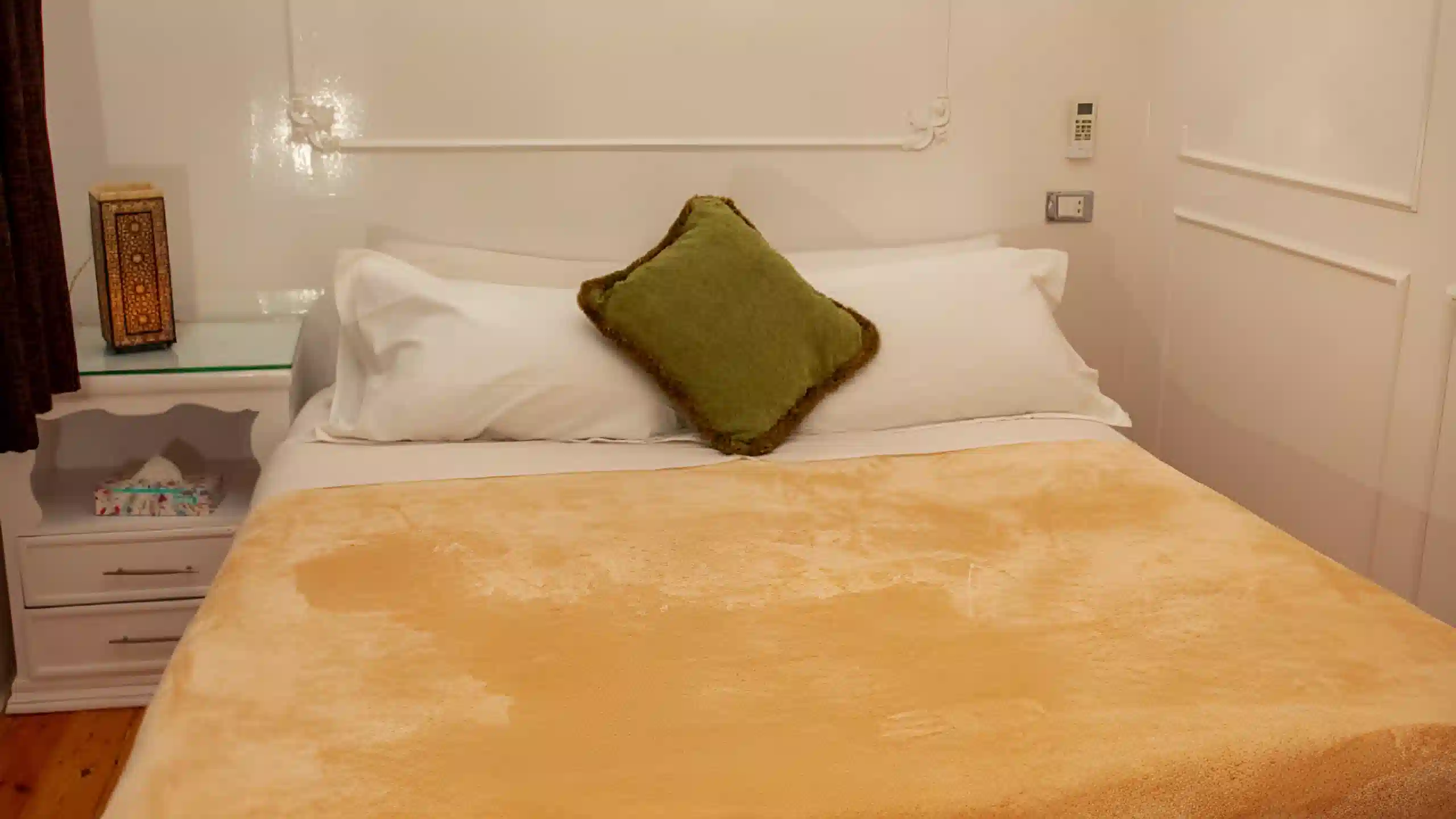 Cozy double bed with golden blanket, green cushion, and oriental bedside lamp in a white-walled room.