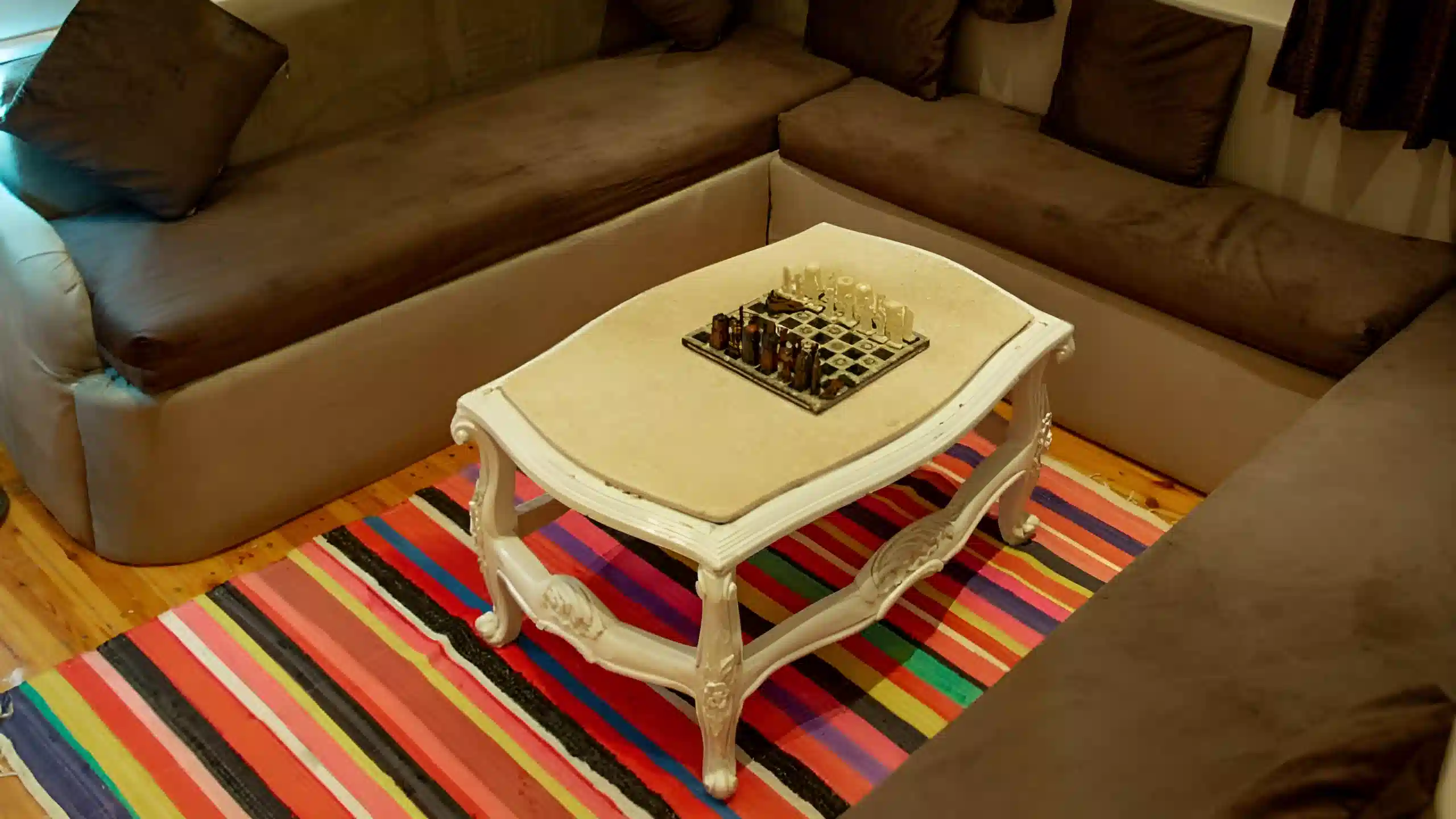 Cozy lounge area with brown corner sofa, vintage white coffee table, colorful striped rug, and a chessboard.