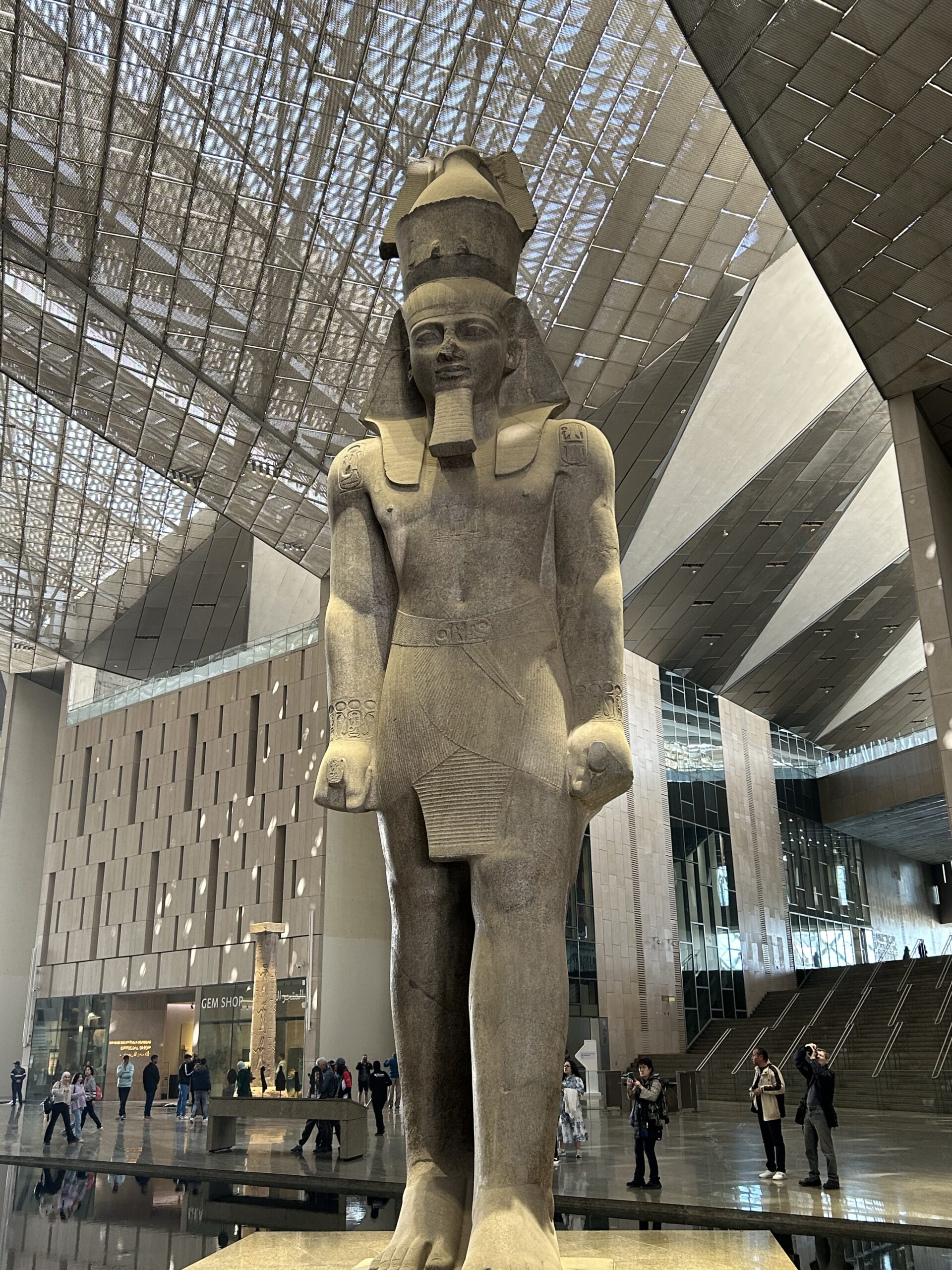 A colossal statue of an ancient Egyptian pharaoh, likely Ramses II, stands proudly in the Grand Egyptian Museum’s vast atrium, surrounded by visitors and modern architectural elements.