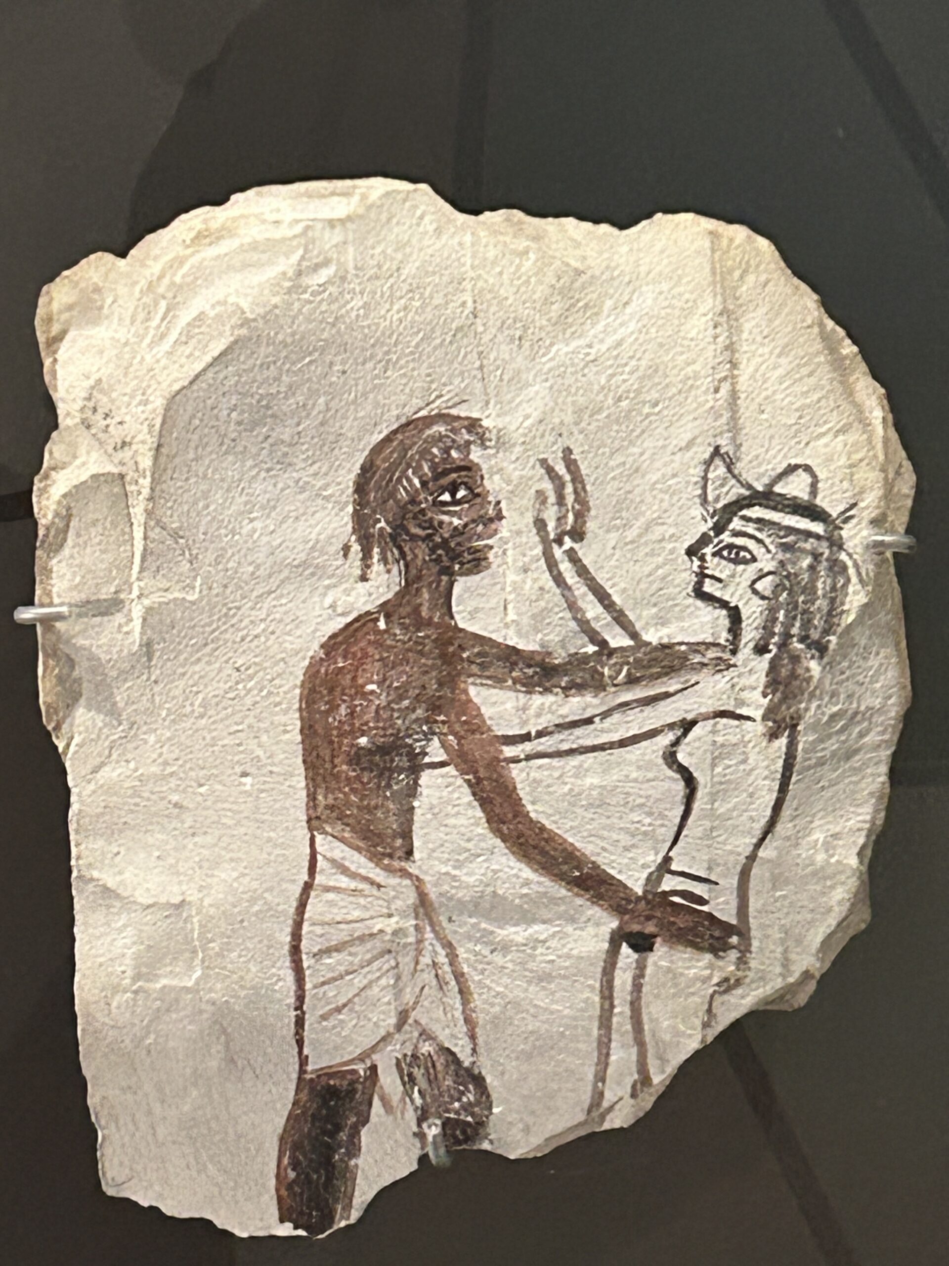 A painted limestone shard from ancient Egypt showing a man in a white kilt facing a female figure with a feline head, likely a humorous or satirical depiction, displayed in the Grand Egyptian Museum.