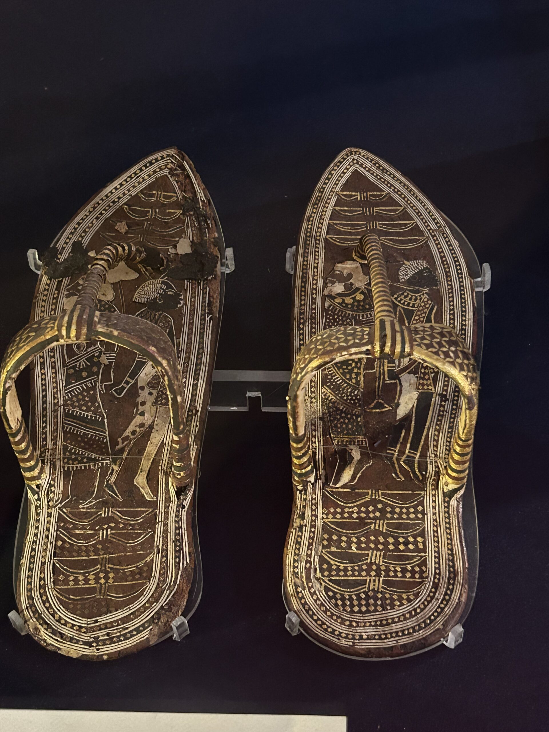 A pair of ornate wooden sandals that belonged to Pharaoh Tutankhamun, intricately decorated with gold-painted scenes and symbolic motifs, displayed in a museum case.