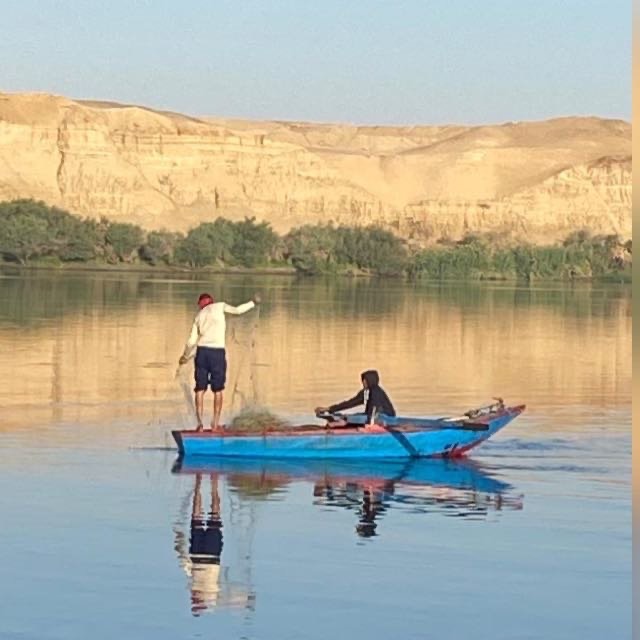 A father and his son in a small blue fishing boat on the calm Nile River—while the father casts a net, the son rows quietly. Golden cliffs and green trees reflect softly in the still water.