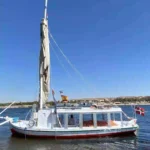 A white traditional Egyptian felucca with multiple flags, moored on the Nile River under a clear blue sky.