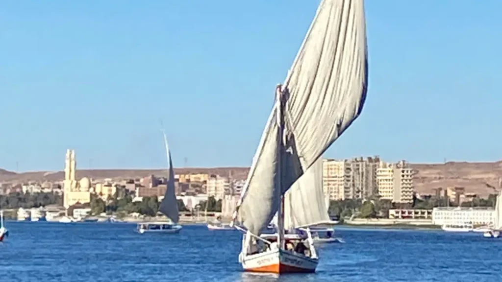 Traditional felucca sailing on the Nile near Aswan with city buildings along the riverbank