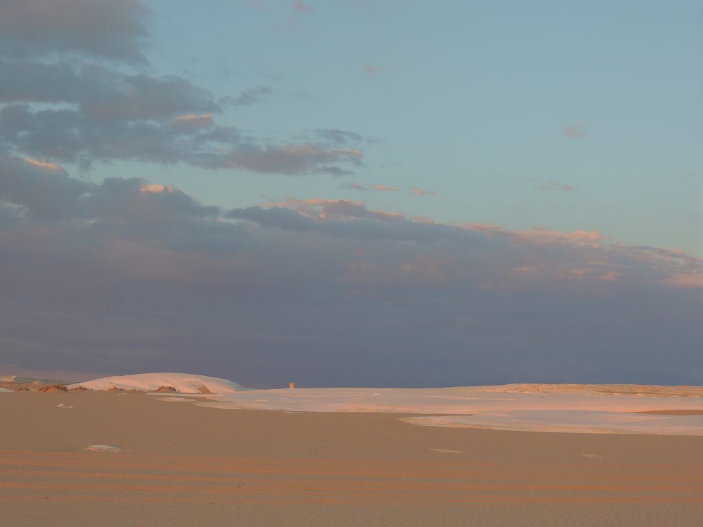 Wide desert landscape with soft sand dunes and white limestone plateau under dramatic cloudy sky at sunset in Egypt’s White Desert
